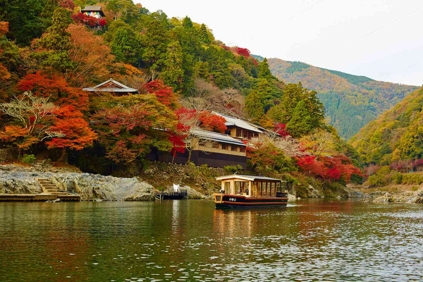 Hotel con vista sul fiume, attraversato da un imbarcazione in autunno con foliage di colore rosso-arancio. In Giappone