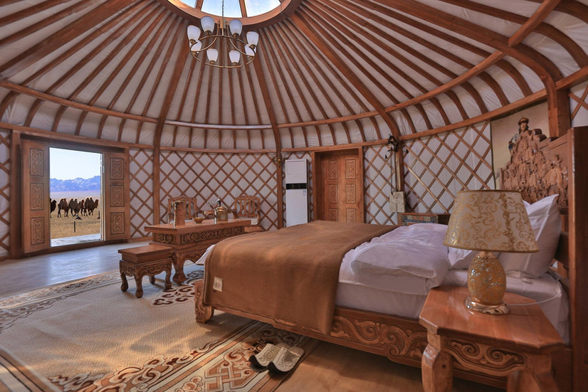 Interno di una elegante yurta con struttura e arredi in legno intarsiato con tappeti tradizionali, fuori dalla porta principale vista su gruppo di cammelli