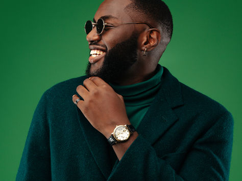Coily haired Man in a dark green blazer and round sunglasses, smiling against a matching green background. He wears a watch and ring, exuding joy.
