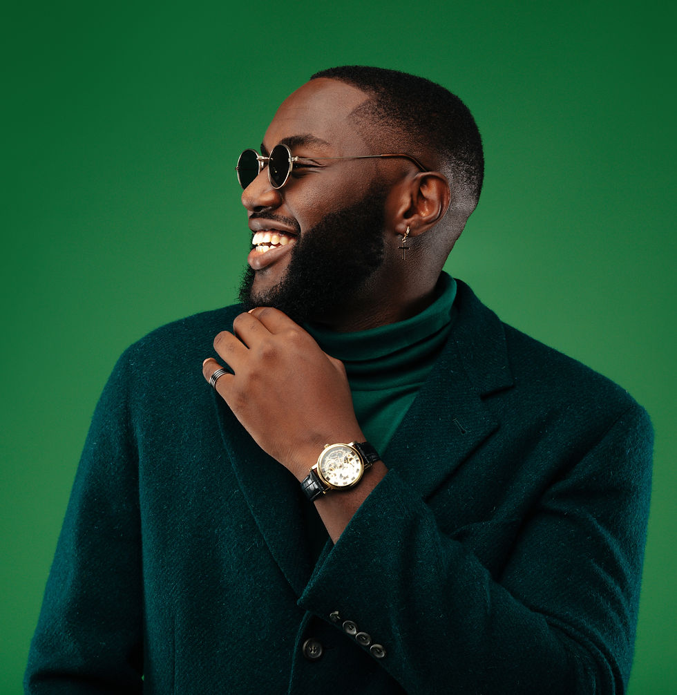 Coily haired Man in a dark green blazer and round sunglasses, smiling against a matching green background. He wears a watch and ring, exuding joy.