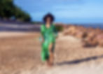 Woman in green dress walking on a sunny beach. Trees line the left, rocky shore to the right. Blue sky and ocean in the background.