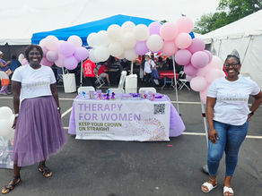 Two smiling women stand at a balloon-decorated Therapy for Women booth under a tent at an outdoor fair.