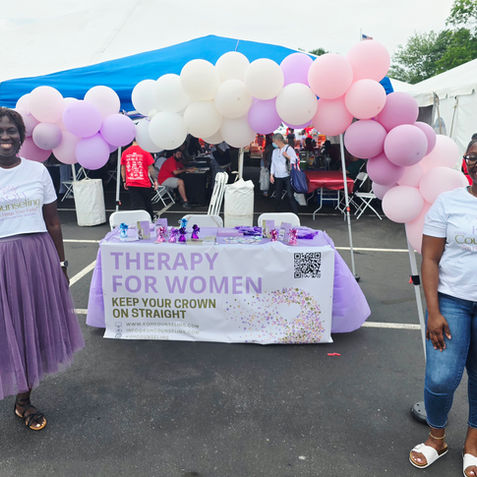 Two smiling women stand at a balloon-decorated Therapy for Women booth under a tent at an outdoor fair.