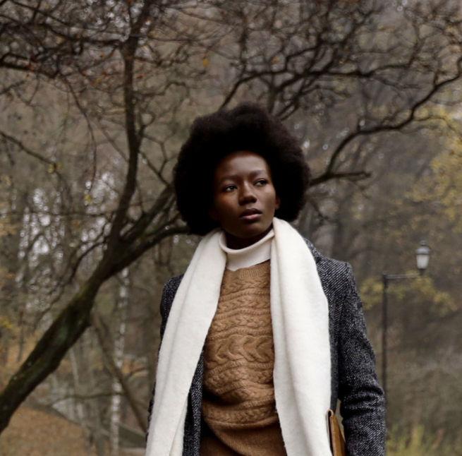 Woman in a gray coat and white scarf stands in a bare park, looking left with a calm, thoughtful expression.