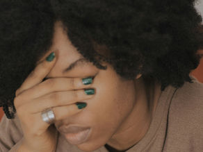 Person with curly hair covers face with hand, showing green nail polish. Brown shirt, neutral background. Mood appears tired or stressed.