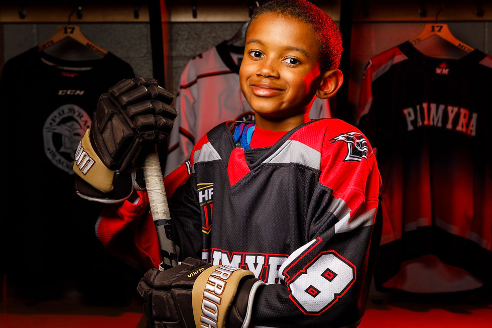 A youth hockey player smiles and holds his stick in a creatively lit locker room.