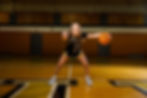 A women's basketball player points at the camera while dribbling inside a gymnasium.