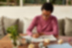 A person in a pink shirt writes in a notebook at a wooden table, surrounded by crumpled papers and a plant, in a cozy room with cushions.