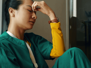 A person in green scrubs, sitting with eyes closed, rests their head in hand, conveying exhaustion. Background is neutral with a potted plant.