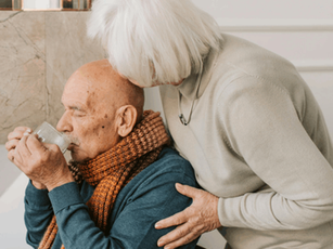 Elderly man in blue sweater and brown scarf drinks from cup. Woman in beige sweater leans over, showing care. Background with marble wall.