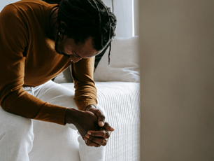 Man sitting on bed, head down, hands clasped. Wearing brown shirt, white pants. Neutral-toned room. Mood appears contemplative.
