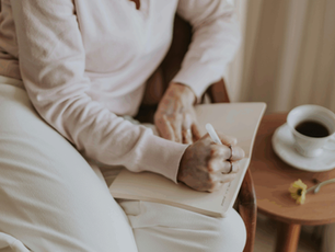 Person writing in a notebook on their lap, sitting in a cozy chair. A cup of coffee and a yellow flower are on a wooden table nearby.