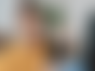 Young woman in a yellow sweater smiles and waves at a phone during a video call, sitting on a couch with plants in the background.