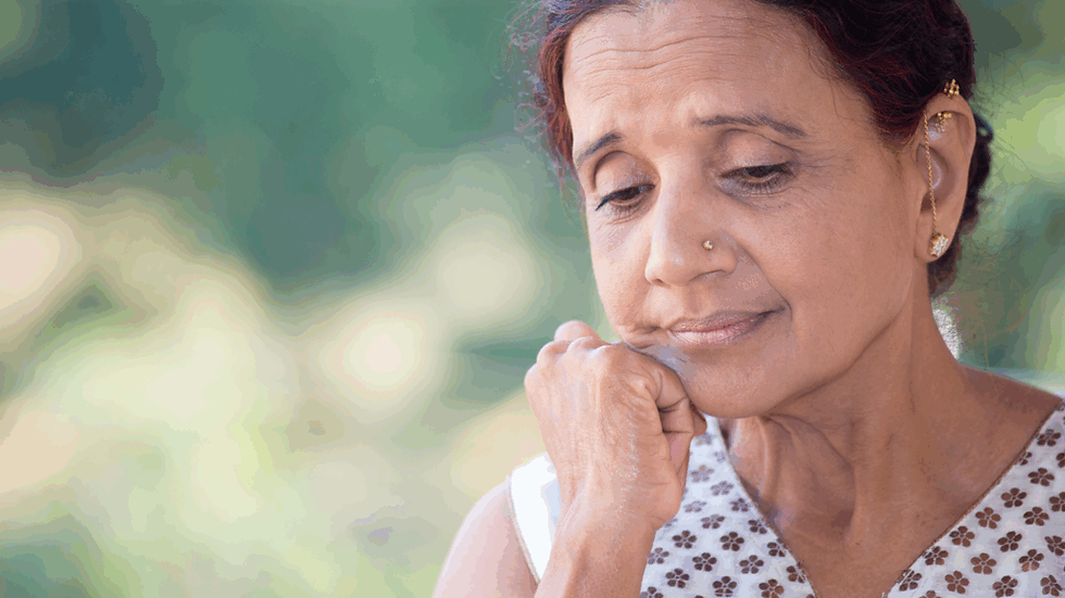 Elderly woman with chin on hand, appearing thoughtful, wears a patterned top and gold earrings, against a blurred green background.