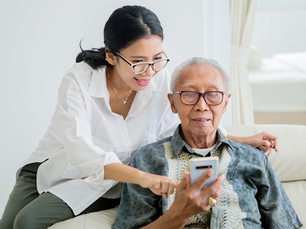 Woman in white shirt helps elderly man with a phone. They're sitting on a cream sofa in a bright room, both smiling and focused.