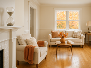 Warm, freshly cleaned living room decorated for Thanksgiving with beige furniture, autumn pillows, pumpkins, fall floral arrangement, and an hourglass on the mantel symbolizing ‘time matters,’ sunlight shining through windows with colorful fall leaves outside.