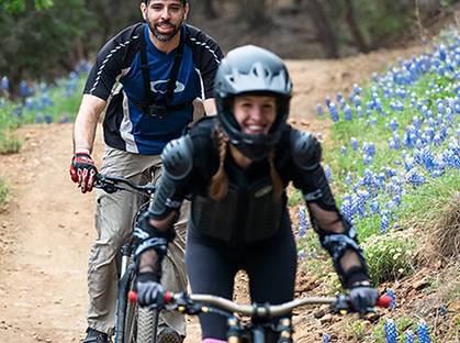 Two glamping guests mountain biking downhill on a dirt path lined with bluebonnets in Marble Falls, TX