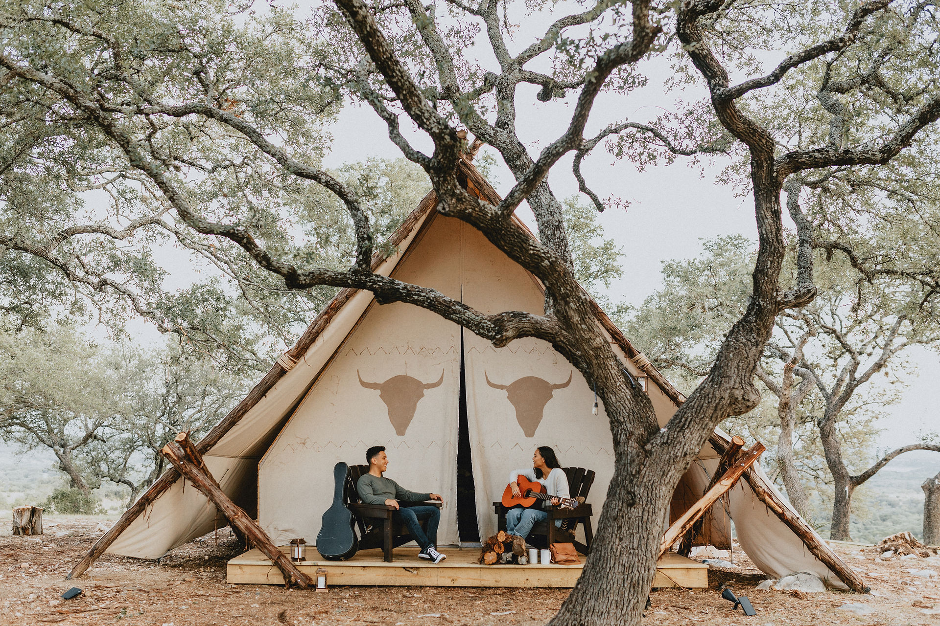 Gorgeous glamping near Marble Falls, Texas. Talula Mesa glamping tent or dome, with a couple playing guitar and relaxing in Adirondack chairs on the front porch of their glamping tent, surrounded by live oak trees and nature.