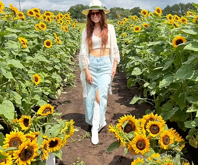 Woman picking buckets of sunflowers in a sunflower field near Austin, TX. She is wearing sunglasses, a sun hat, tank top, long denim skirt, and white cowgirl boots.