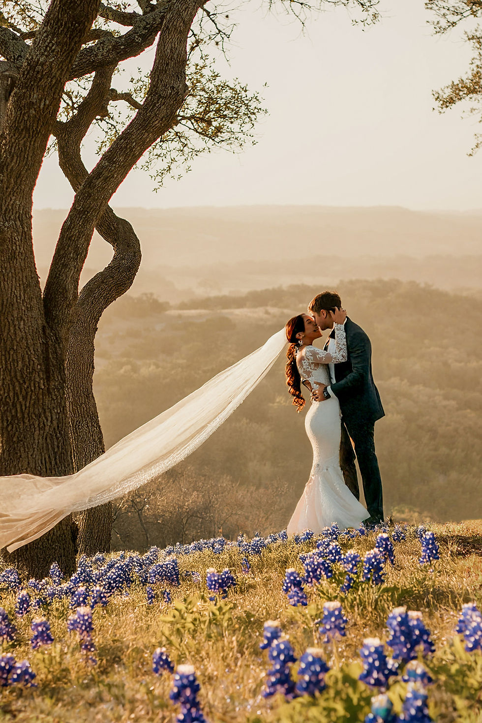 A bride and groom having a Texas Bluebonnet wedding at Talula Mesa Glamping Resort in the Texas hill country. The couple is embracing, looking at each other and standing on the top of a flat hill covered in bluebonnets with green and golden rolling hills behind them. The bride's veil is blowing gently in the wind. Overcast sky. Romantic, ethereal atmosphere.