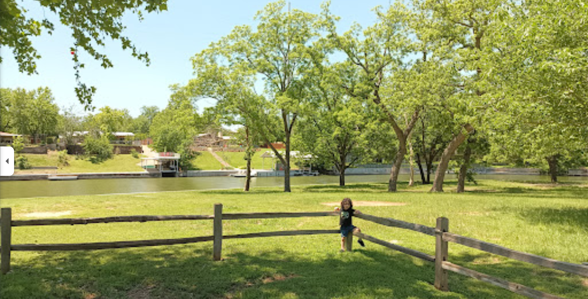Park with grass, trees, and child climbing on a wooden fence in Marble Falls, Tx near Talula Mesa Glamping Resort.