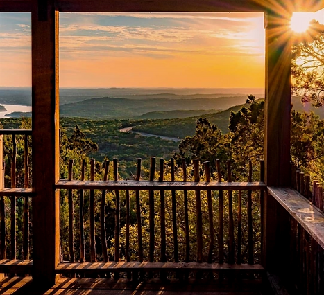 View of the Texas Hill country, Lake Travis, and Marble Falls, TX near Austin through the Sunset Deck at the Balcones Canyonland National Wildlife Refuge. The sunset deck is a wooden hut, and the view is looking out from the hut over the hill country at sunset, over the green hills framed by an orange sky.
