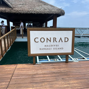 Conrad Maldives Rangali Island sign in front of thatched roof building, ocean background.