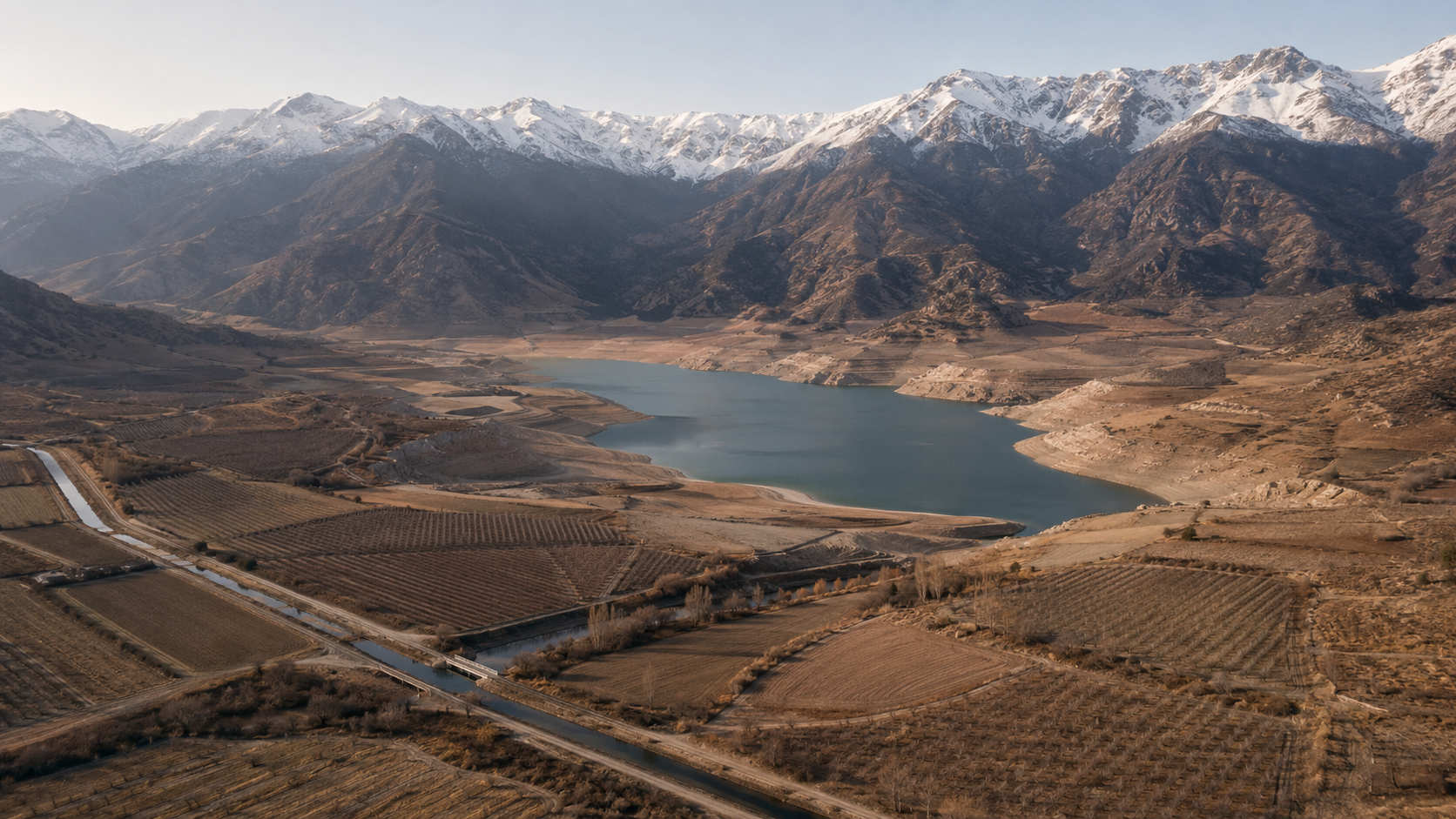 Vista aérea editorial de un valle andino chileno en pleno invierno temprano — cordillera con primera nieve a cota media, campos en descanso y embalse parcial en el valle