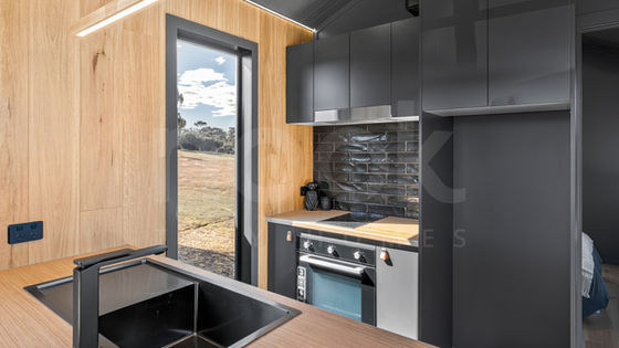alternate kitchen view of the new voyager series tiny home on wheels THOW by Nook tiny homes - beautiful modern finish with feature timber wood panelling on walls and timber flooring, luxury kitchen and appliances, large format glass windows