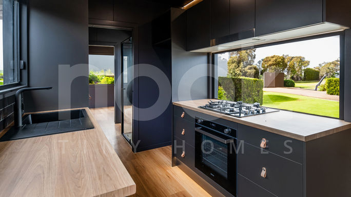 A different angle of the modern kitchen in a 40ft container home by Nook Tiny Homes. This view highlights the built-in gas stove and oven, black cabinetry, light wood countertops, and large windows. The light wood flooring continues to add warmth to the sleek design.