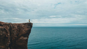 Lone hiker in a yellow jacket stands on a rocky cliff above the calm blue ocean under a cloudy sky.