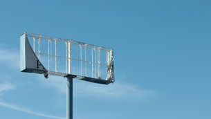 Damaged empty billboard frame on a pole against a clear blue sky, with a partial P visible on the left.