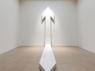 Single white marble arrow sculpture on a bare gallery floor, pointing forward