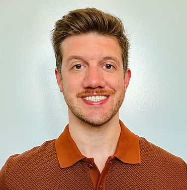Dr. Tyler Degener with brown shirt and light hair smiling in front of white wall.
