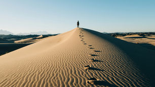 Lone hiker stands atop a sand dune, footprints trailing behind in a vast desert under a clear blue sky.