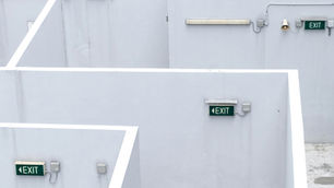 White stairwell wall with three green EXIT signs, a fluorescent light and alarm fixtures, bright geometric shadows.