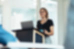 A business professional at a podium with windows behind her doing a presentation
