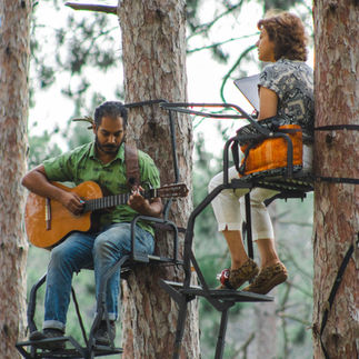 Musicians Shinjan Sengupta and Ritika Ganguly perform. Shinjan plays the guitar. He has brown skin and dark hair and is wearing a green shirt and jeans. Ritika has shoulder-length brown hair and brown skin and sings, lots of emotion on her face. Both performers sit in deer stands in Belwin's red pine grove.