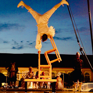 Ein Akrobat führt bei Dämmerung eine beeindruckende Handstand-Performance auf gestapelten Stühlen während einer Outdoor-Show vor historischer Kulisse vor. Perfekt für beeindruckende Märchen Shows und Gala Dinner-Events.