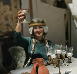 Gertrud von Haldensleben Rede und historischer Walk Act, historische Frau Herzogin aus dem 11. und 12. Jhdt., sitzende Dame in früh mittelalterlichem blau grünem Gewand mit langen blonden geflochtenen Zöpfen und Schapel mit Schleier, lächelt und prostet mit Tonkelch in Richtung Kamera