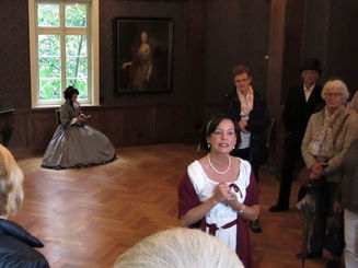 Annette von Droste-Hülshoff, 19. Jhdt. Gedicht Lesung Rezitation und Vortrag, historische Führung im Herrenhaus, im Vordergrund Museumsleitung Jutta Törnig-Struck bei Führung, um sie herum interessiertes Publikum, im Hintergrund historische Frau mit Haube, historischer Lockenfrisur und grauem Biedermeier Kleid / Kostüm, sitzt vor Fenster unter einem Gemälde an der Wand, liest aus kleinem Gedichtband, historischer Walk Act in Gut Rödinghausen