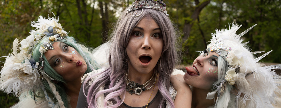 Zauberschule Geist Feen: junge Frau mit grauer Perücke und Perlenkette schaut Stunden in die Kamera, rechts und links von ihr zwei Frauen mit Glitzer schminke, weiß-bläulichen Haaren und aufwendigem weißem Feder- und Glitzer Haarschmuck, schauen ihr über die Schulter, eine streckt die Zunge aus. immersives Event