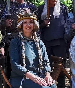 Gertrud von Haldensleben Rede und historischer Walk Act, historische Frau Herzogin aus dem 11. und 12. Jhdt., sitzende Dame in früh mittelalterlichem blau grünem Gewand mit langen blonden geflochtenen Zöpfen und Schapel mit Schleier, lächelt