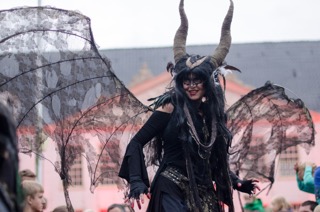 fantastischer Walk Act Dämon: Frau auf Stelzen mit großen geschwundenen Hörnern und großen halb durchsichtigen, zerfetzten Fledermaus - Dämonenflügeln, mit schwarzer Maske mit Federn, Stelzen Walk Act