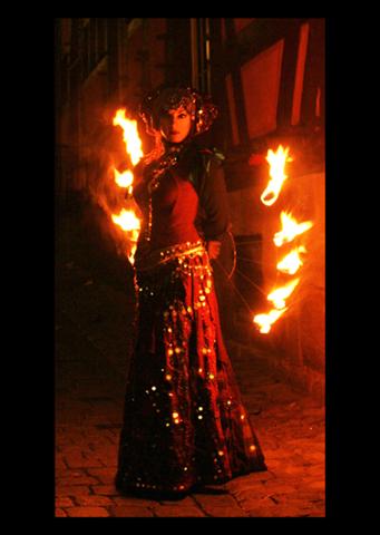 Frau mit fantasievoll dunklem glitzerndem Gewand im Tribal Stil und Dreadlock Haarschmuck hält Feuerfächer hinter dem Rücken verschränkt