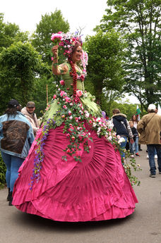Eine Künstlerin auf Stelzen in einem opulenten Blumenkleid verzaubert als Walk Act das Publikum. Das farbenfrohe Kostüm mit Frühlingsblüten, Ranken und Blätterkrone bringt den Frühling auf stilvolle und poetische Weise zum Leben.