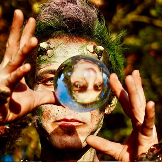 Ein fantasievoll geschminkter Darsteller in grüner Waldkostümierung hält eine Kristallkugel, die sein Gesicht auf faszinierende Weise spiegelt. Perfekt für mystische Walk Acts, Shows und exklusive Gala Dinner-Events.