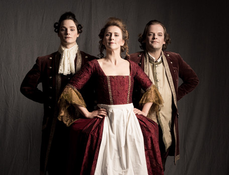 Rokoko Theater Dienerschaft. Drei Diener, vorne eine blonde Zofe in rotem Rokoko Gewand und Schürze, mit Händen auf den Hüften, rechts und links dahinter zwei Lakaien in mauvefarbenen Livreen, Hände hinter dem Rücken