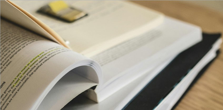 Four books stacked on top of a wooden desk. Top of pile has a yellow sticky tab packet sitting on top.