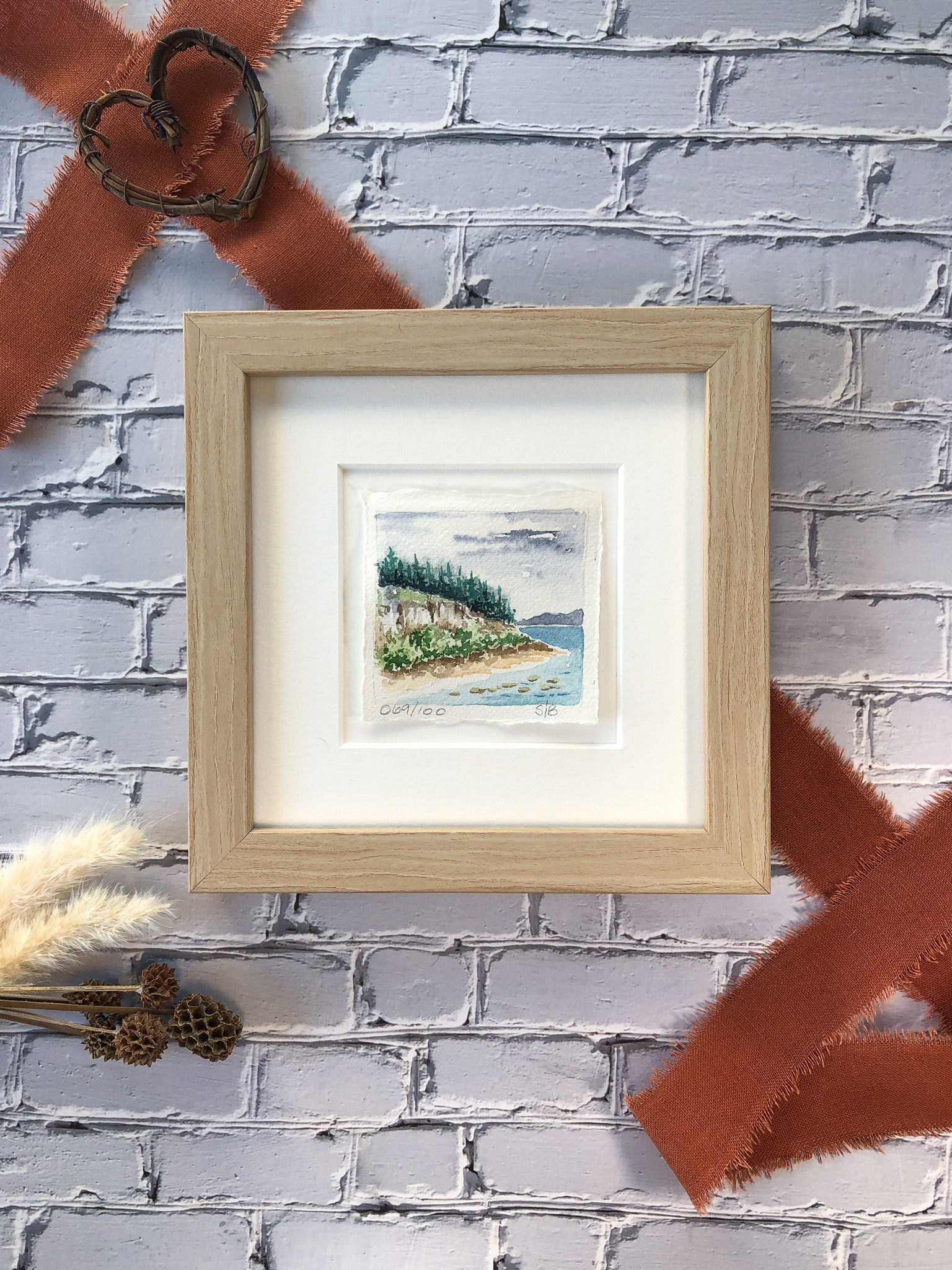 watercolor painting of rocky shore of lake, pine trees, framed with rust ribbon accent